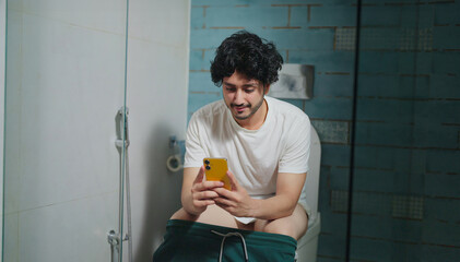 Indian Asian active young adult gen z happy man guy sitting on toilet use mobile phone online share daily news teen age male boy hold gadget check social media read typing text bad habit indoor home