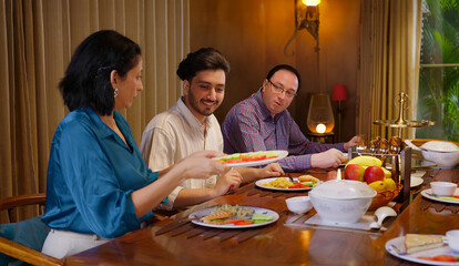 Indian family group sitting around dining table chair enjoy night dinner indoor home happy mom lady serve fresh salad cute kid boy dad hold spoon eat tasty food talk gossip have fun joy spend time