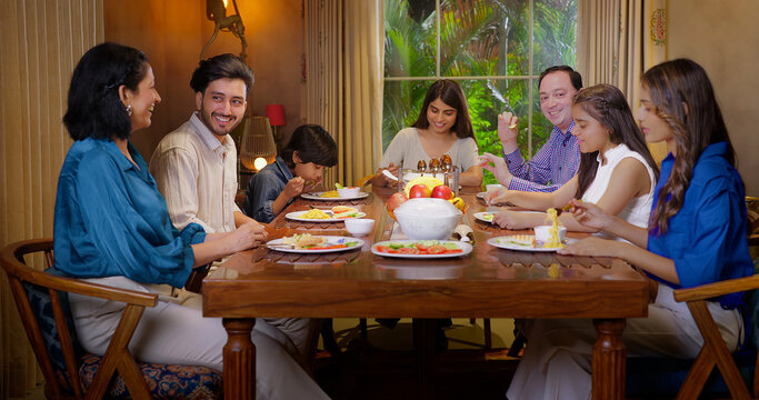 Indian happy family group with cute kid child sitting around dining table chair enjoy night dinner adult dad teen girl young boy talk mom lady serve fresh salad eat tasty food have fun joy spend time - Powered by Adobe