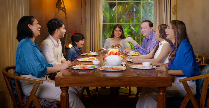 Indian happy family group with cute kid child sitting around dining table chair enjoy night dinner adult dad teen girl young boy talk mom lady serve fresh salad eat tasty food have fun joy spend time