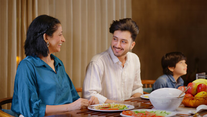 Indian family group sitting around dining table chair enjoy night dinner indoor home. happy mom lady feed with hand serve fresh salad cute kid boy hold spoon eat tasty food have fun joy spend time