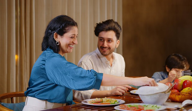 Indian family group sitting around dining table chair enjoy night dinner indoor home. happy mom lady serve fresh salad cute kid boy hold spoon eat tasty food have fun joy spend time - Powered by Adobe