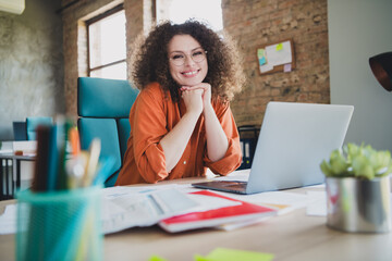 Confident young businesswoman with curly hair smiling at her desk in a modern office setting,...