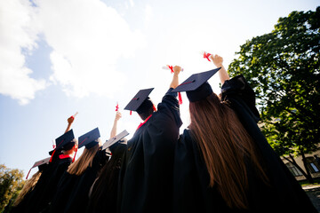 Rear portrait of group students raise diplomas sky mortarboard gown graduation university building...