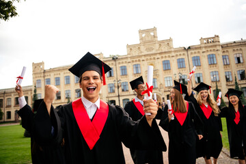 Group of happy graduates celebrating outdoors with diplomas in hand against university backdrop on...