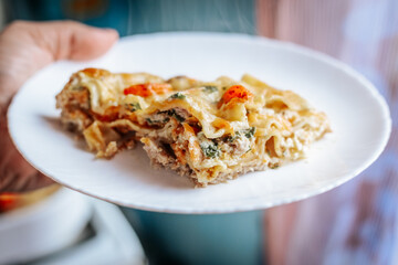 A close-up of a freshly baked slice of lasagna with melted cheese, cherry tomatoes, and herbs on a white plate. The dish appears hot and steaming, ready to be enjoyed.