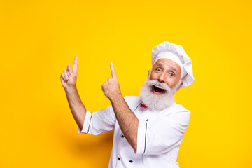 Happy senior chef enthusiastically pointing, wearing uniform on yellow background