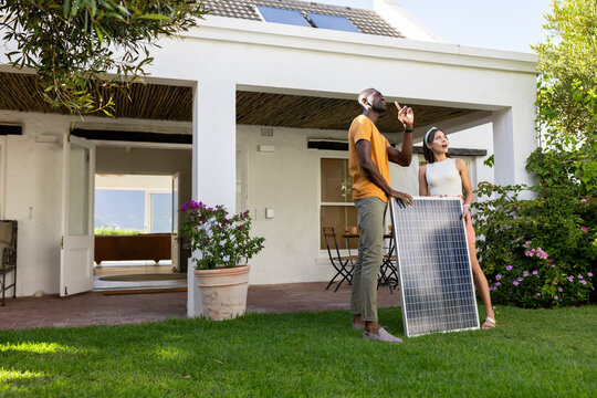 Diverse couple holding solar panel in garden, discussing sustainable energy solutions, copy space