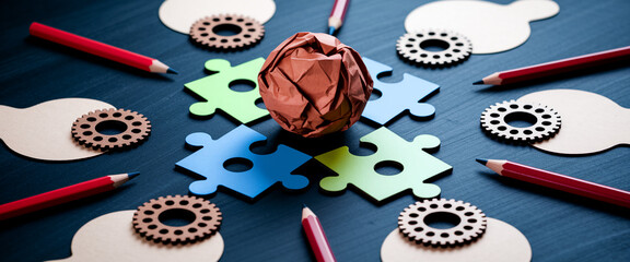 Colored puzzle pieces, crumpled paper ball, and gears surround pencils on a dark surface, symbolizing brainstorming, problem-solving, and creative process