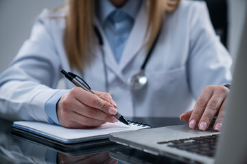 Fototapeta premium Woman doctor in white coat sitting working at desk in the clinic office room, using laptop, working with medical paper documents, writing in notebook.