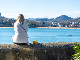 Woman sitting with her back to a wall in front of the touristy La Concha beach in San Sebas on a sunny day enjoying the good weather.