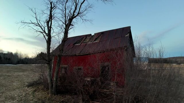 A rustic red barn with a damaged roof stands alone in a field, surrounded by trees and sparse vegetation as evening approaches, casting a serene atmosphere.
