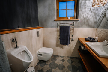 A stylish bathroom with a modern design, featuring a wall-mounted toilet, urinal, wooden countertop, towel warmer, and soft lighting for a cozy atmosphere..