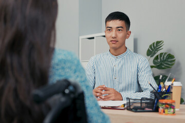 A professional job interview unfolds in an office, where a smiling male boss of Asian-Korean beauty asks questions to a woman who uses a wheelchair. She sits opposite him, answering with confidence.
