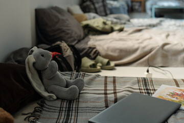 Grey soft toy rabbit sitting in front of laptop and book of comics on warm checkered plaid covering...