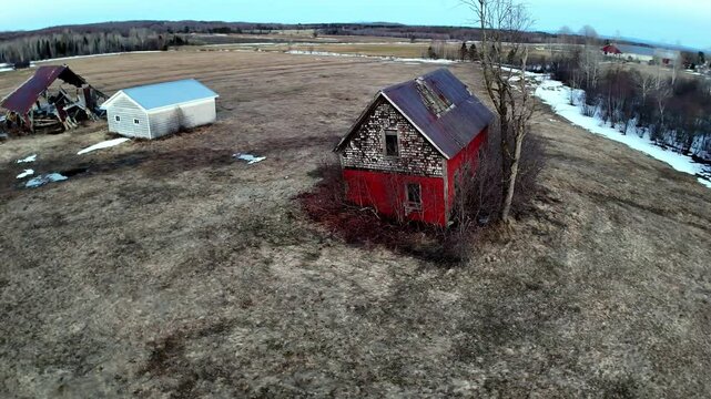 An abandoned red house stands alone in a sprawling rural landscape with patches of snow. Surrounding fields stretch out towards the horizon, creating a serene yet lonely atmosphere.