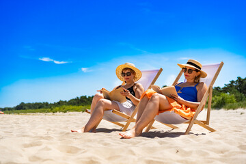 Summer holidays on beach with book. Two mature women reading books sitting on deckchairs on white sandy beach in summertime	