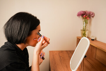 Applying mascara to her brows, a woman admires her reflection in a hand-held mirror. Her short...