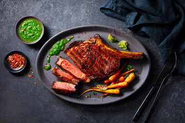Steak tomahawk on a bone with roasted carrots and chimichurri sauce on a plate. Dark background. Close up. Top view.