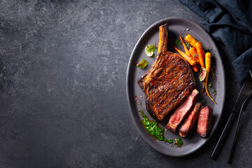 Beef tomahawk steak on a bone with roasted colorful carrots and chimichurri sauce on a plate. Dark...