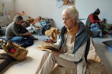 Senior female refugee reading book while sitting on couchette in crisis camp for homeless people against guy and mature woman