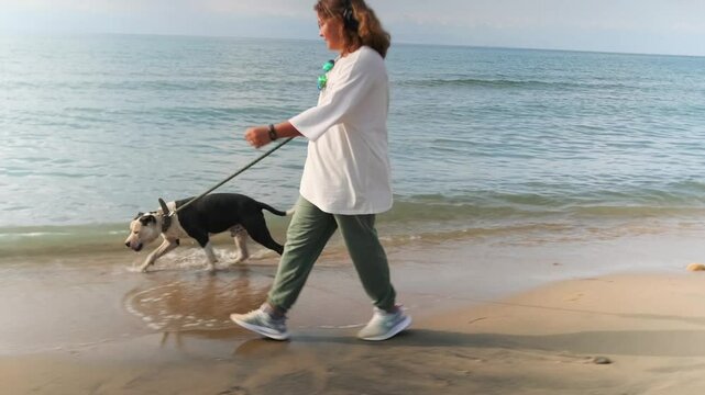 A teenage girl wearing headphones walks along the sandy lake shore, leading a pit bull on a leash, picking up the pace and running as the dog joyfully races ahead, paws brushing the lapping waves.