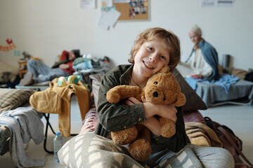 Cute smiling little boy with brown teddybear sitting on couchette in front of camera in camp for...