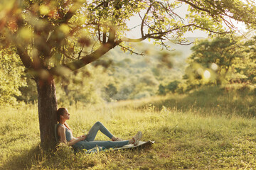 Young woman enjoying a peaceful moment under a tree, wearing casual summer clothing in a serene green landscape, evoking calmness and tranquility