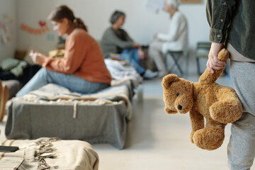 Hand of little boy in black denim jacket and grey sweatpants holding brown teddybear while standing...