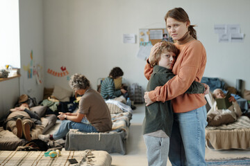 Miserable young woman in casual attire embracing her youthful son and both looking at camera while...