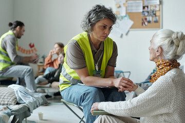 Female volunteer sitting in front of senior refugee and holding her by hand while listening to...