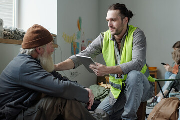 Young male volunteer touching shoulder of senior refugee and comforting him while entering personal...