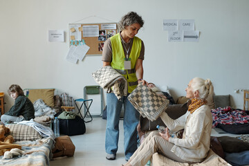 Smiling mature female volunteer in uniform passing folded blanket to senior woman sitting on...