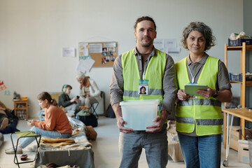 Young and mature volunteers in reflective jackets looking at camera while working in refugee camp...