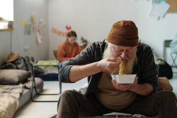 Hungry senior male refugee with long grey beard eating noodles from bowl in crisis camp while...