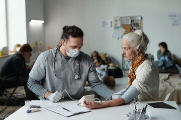 Man in medical scrubs and protective mask using tonometer for measuring blood pressure of senior female settler of crisis camp