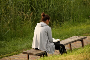 A young girl in a gray sports jacket sits on a bench in the park and reads a book. Intellectual...