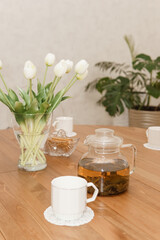 A peaceful arrangement featuring a glass teapot, cups, and a vase of white tulips on a wooden table, evoking warmth, simplicity, and serenity in the decor.