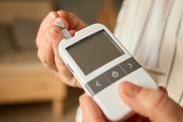 A person is using a glucometer to check their blood sugar levels. The device is held carefully in their hands, with a background of a comfortable living room