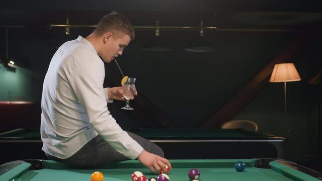 Player in white shirt and checkered trousers sits on green pool table, sipping lemon drink while rolling billiard ball. Stylish billiards lounge with ambient lighting creates a relaxed atmosphere