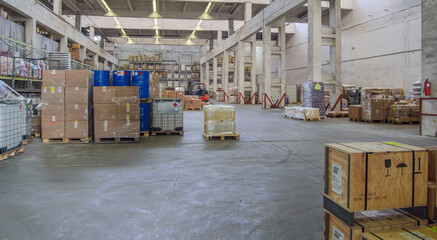 The spacious warehouse interior is filled with boxes and barrels placed on pallets. High ceilings and fluorescent lighting contribute to a well-lit environment, while the various goods are visible.