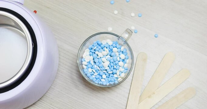 Blue, white wax beads filling glass cup near wooden spatulas and professional wax melter, preparing for precise hair removal procedure