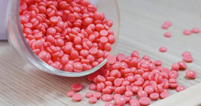 Pink film wax beans cascading from glass jar onto wooden surface, signaling professional depilation preparation in beauty salon setting