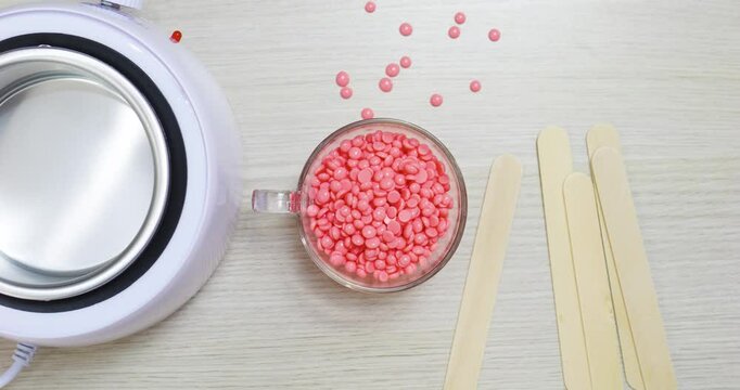 Pink film wax granules beside wooden spatulas near electric wax warmer, preparing professional hair removal treatment on light wooden surface