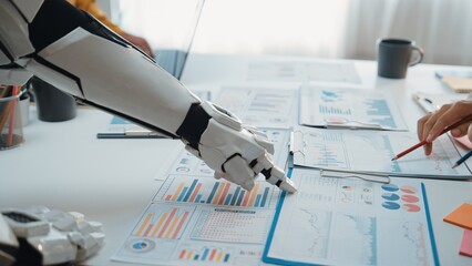 A futuristic robot arm collaborates with a human hand over business reports on an office desk, showcasing the synergy of technology and human effort in data analysis.Noogenesis