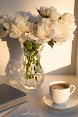 Morning light with coffee and peony flowers. A serene morning setting featuring an elegant bouquet of white peonies in a glass vase, classic white cup of coffee and a closed book. Warm sunlight.