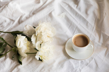 Morning coffee and peony flowers in bed. A tranquil morning routine with a cup of coffee amidst fresh white peonies on a soft linen textile background, conveying a sense of calm, serene, slow living.