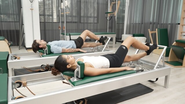 Couple practicing leg exercises on pilates reformer to strengthen core and improve flexibility at gym. Pilates partner doing pelvic bridge pose on reformer for improving lower back muscle. Habituate.
