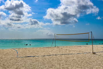 a volleyball net on a sandy beach. turquoise water of the ocean is in the background. several people are swimming in the distance.