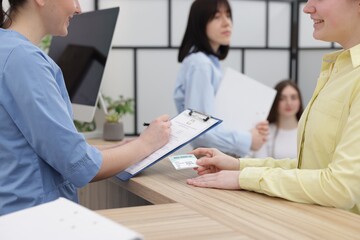 Obraz premium Patient with medical insurance card and receptionist filling document at wooden counter in clinic, closeup
